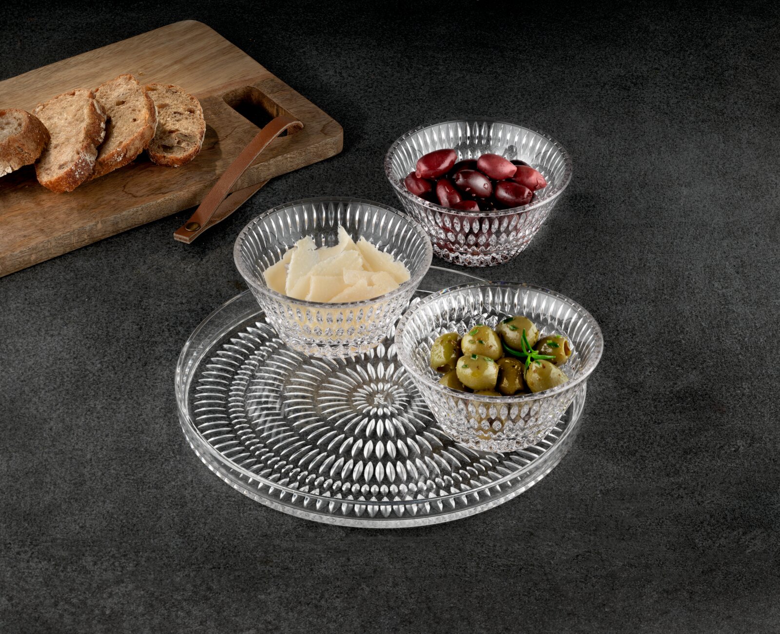 Drie geribbelde glazen schalen met olijven, kaasvlokken en rode olijven op een decoratieve glazen schaal, daarnaast een houten plank met sneetjes brood; perspectief: licht van bovenaf in schuin aanzicht