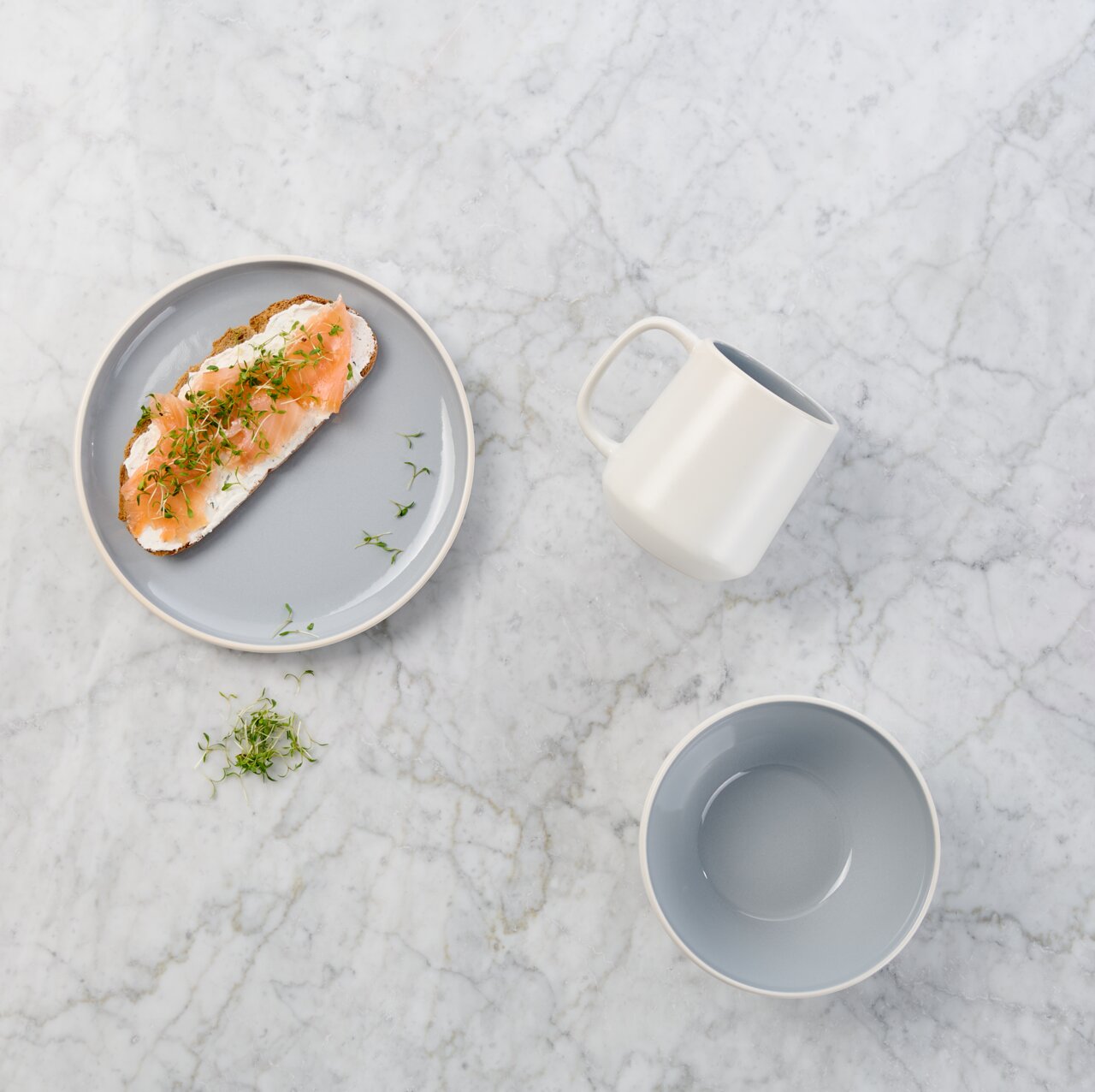Draufsicht auf ein minimalistisches Geschirr-Set auf heller Marmorfläche: grauer Teller mit belegtem Lachsbrötchen, weiße Tasse und graue Schale.
