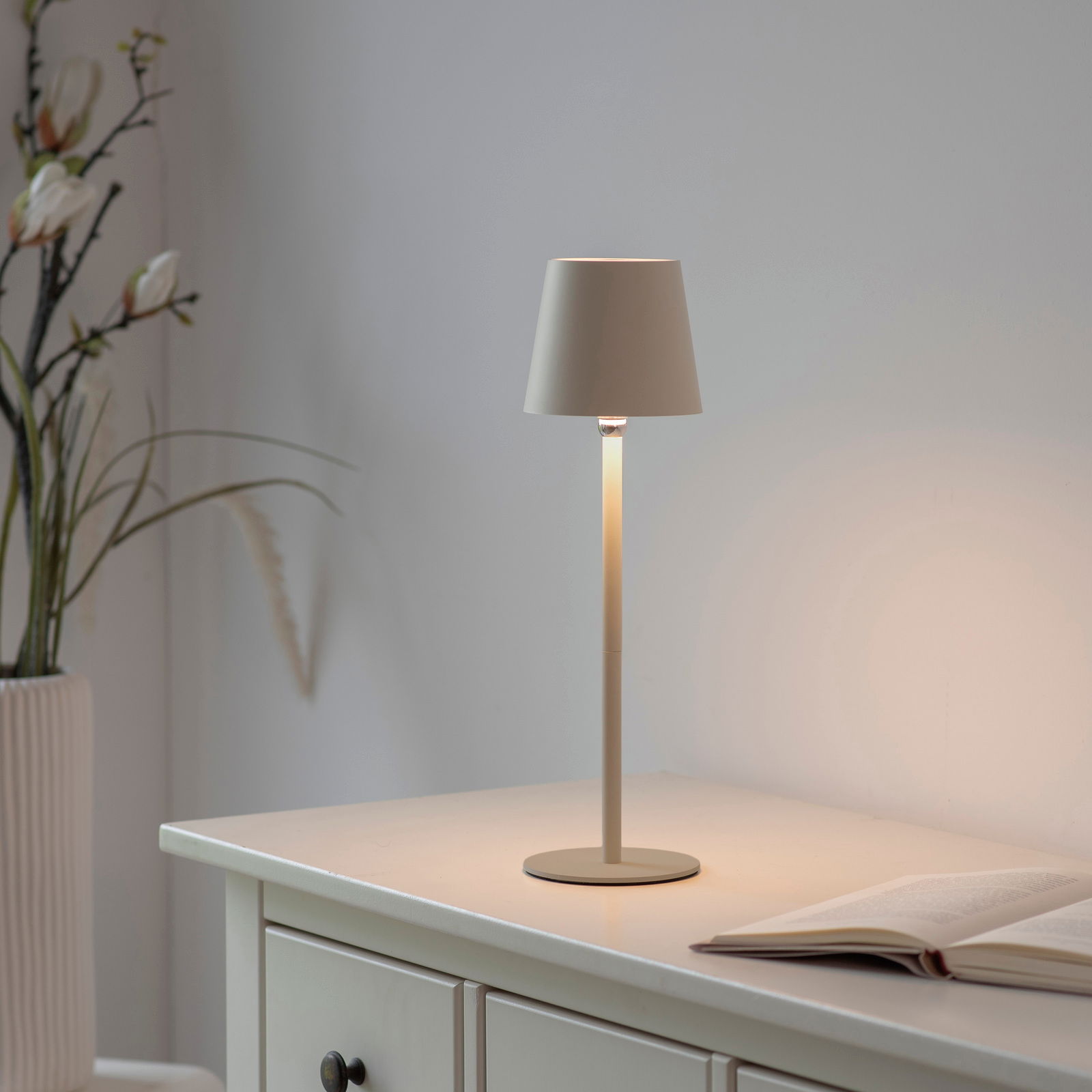 Elegante Akku-Tischleuchte BOTERIA auf einem Tisch, seitliche Perspektive, mit sanftem Lichtschein und dekorativen Pflanzen im Hintergrund.