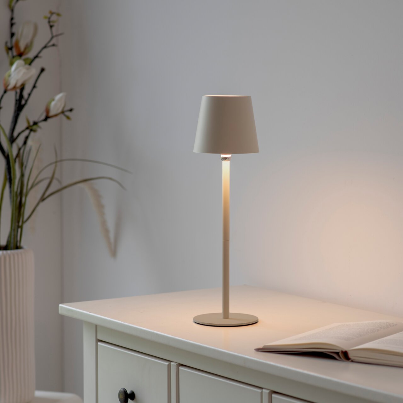 Elegante Akku-Tischleuchte BOTERIA auf einem Tisch, seitliche Perspektive, mit sanftem Lichtschein und dekorativen Pflanzen im Hintergrund.