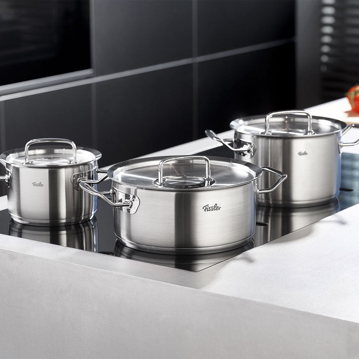 Fissler Kochtopf 28cm ORIGINAL-PROFI COLLECTION 2 Drei Edelstahlkochtöpfe mit Metalldeckeln auf einem modernen Herd, seitliche Perspektive.