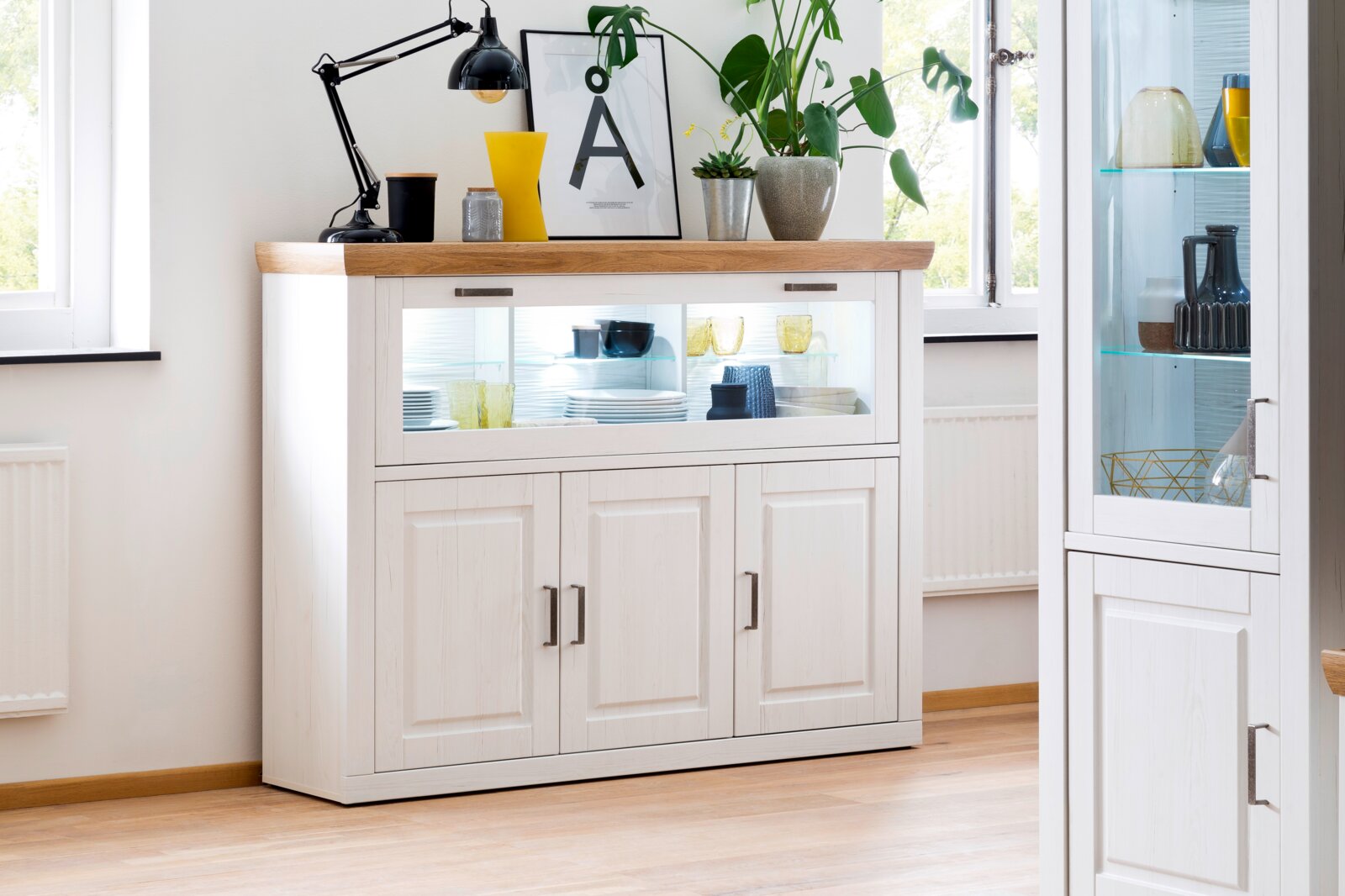 Landscape Highboard BREGENZ Weiße Anrichte im Landhausstil mit Holzplatte und beleuchtetem Glas-Schiebefach, frontal in leicht erhöhter Perspektive fotografiert, dekoriert mit Lampe, Pflanzen und Geschirr.