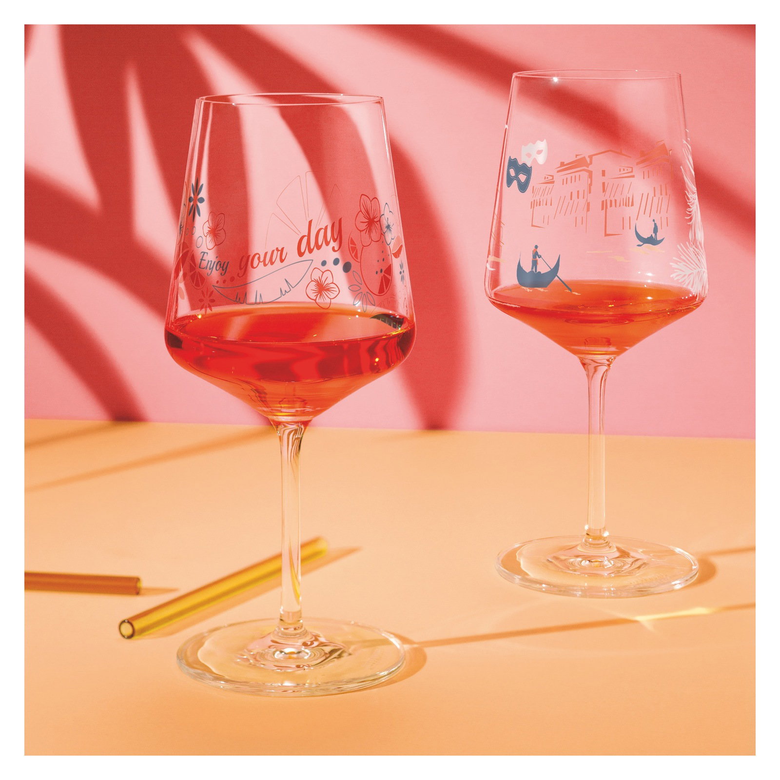 Zwei Weingläser mit dekorativen Motiven und rotem Getränk, auf einem Tisch mit rosa Hintergrund, seitliche Perspektive.