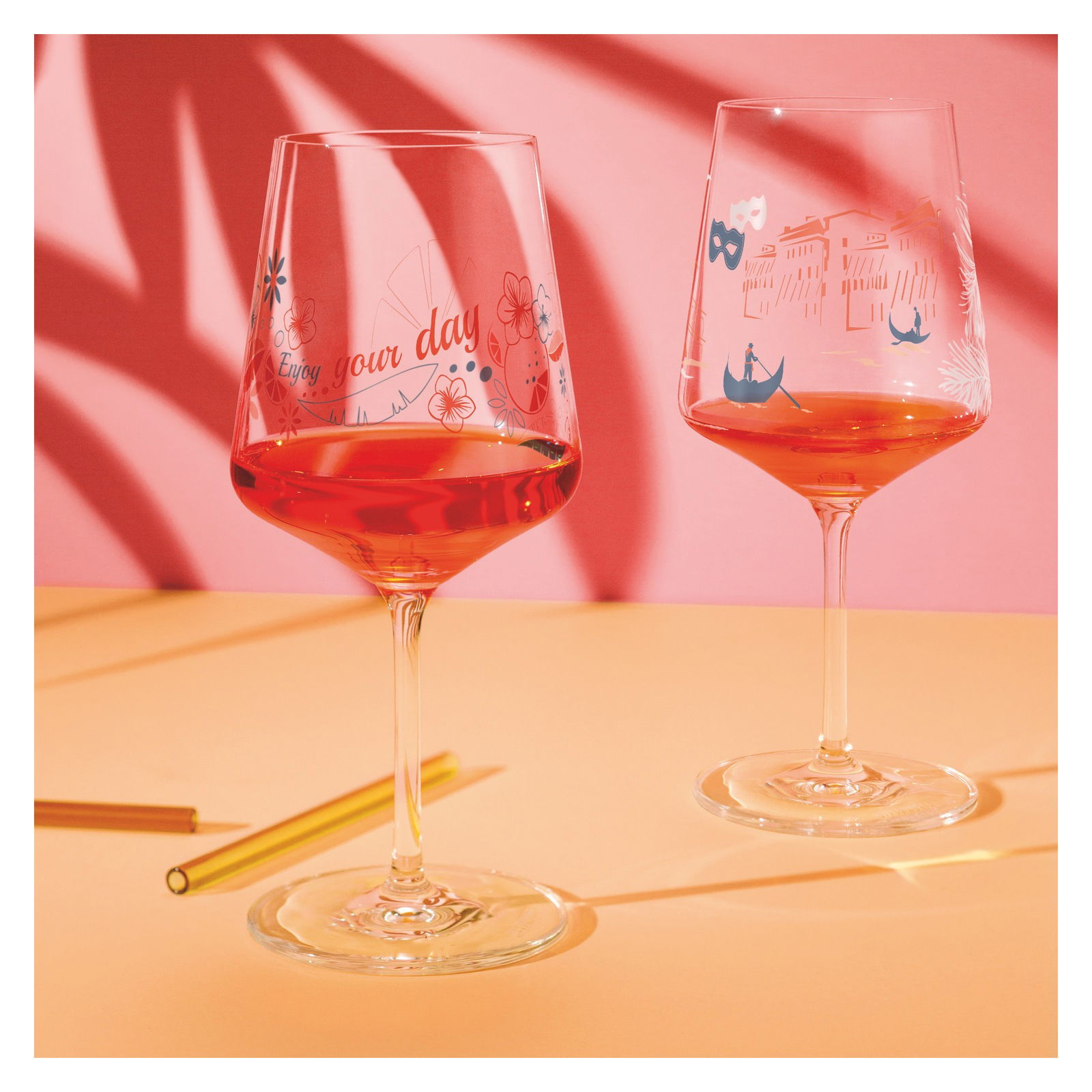 Zwei Weingläser mit dekorativen Motiven und rotem Getränk, auf einem Tisch mit rosa Hintergrund, seitliche Perspektive.