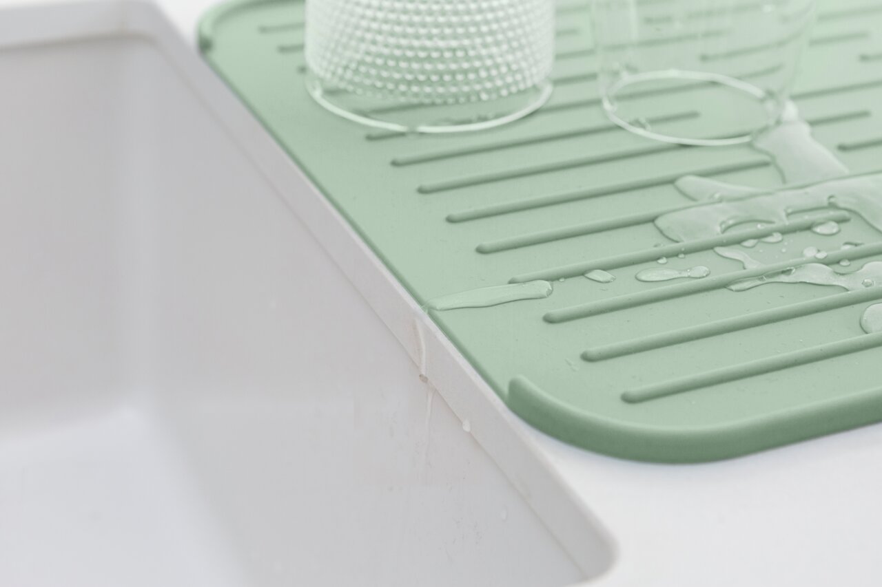 Grüne Silikon Abtropfmatte neben einem Waschbecken, mit Wasser und Gläsern darauf, aus seitlicher Perspektive.