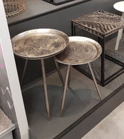 Zwei runde Beistelltische mit antik-silberfarbenen Tablettplatten und schlanken Dreibein-Metallgestellen, schräg von vorne in einem Ausstellungsraum fotografiert