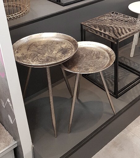 Zwei runde Beistelltische mit antik-silberfarbenen Tablettplatten und schlanken Dreibein-Metallgestellen, schräg von vorne in einem Ausstellungsraum fotografiert