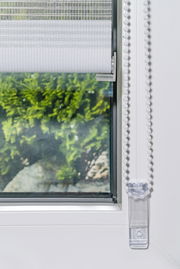 Doppelrollo mit Blende, teilweise heruntergelassen, an einem Fenster montiert, mit Blick auf einen grünen Garten im Hintergrund.