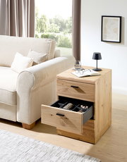 Rollcontainer aus Holz mit zwei Schubladen, eine geöffnet mit Fächern, neben einem Sofa in einem hellen Wohnzimmer, seitliche Perspektive.