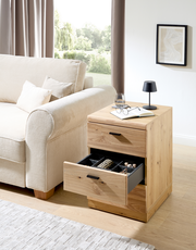 Rollcontainer aus Holz mit zwei Schubladen, eine geöffnet mit Fächern, neben einem Sofa in einem hellen Wohnzimmer, seitliche Perspektive.