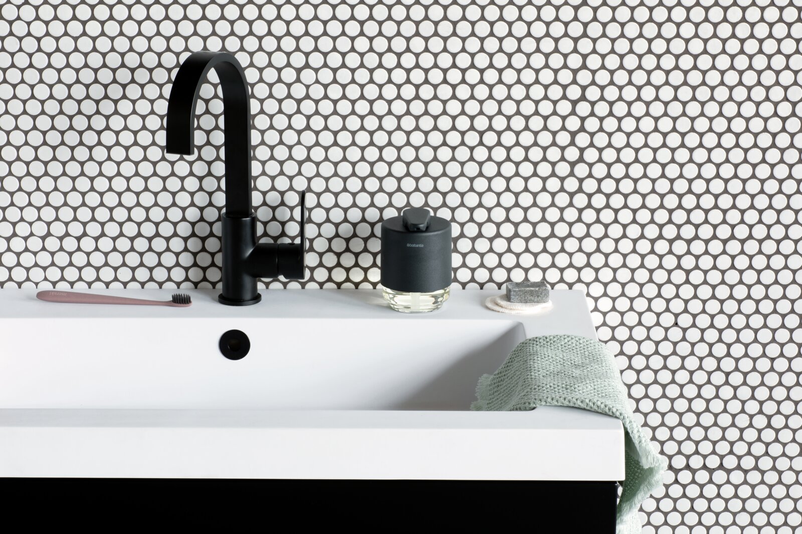 Moderner Seifenspender auf einem Waschbecken mit schwarzem Wasserhahn, im Hintergrund eine Wand mit kreisförmigem Muster, seitliche Perspektive.