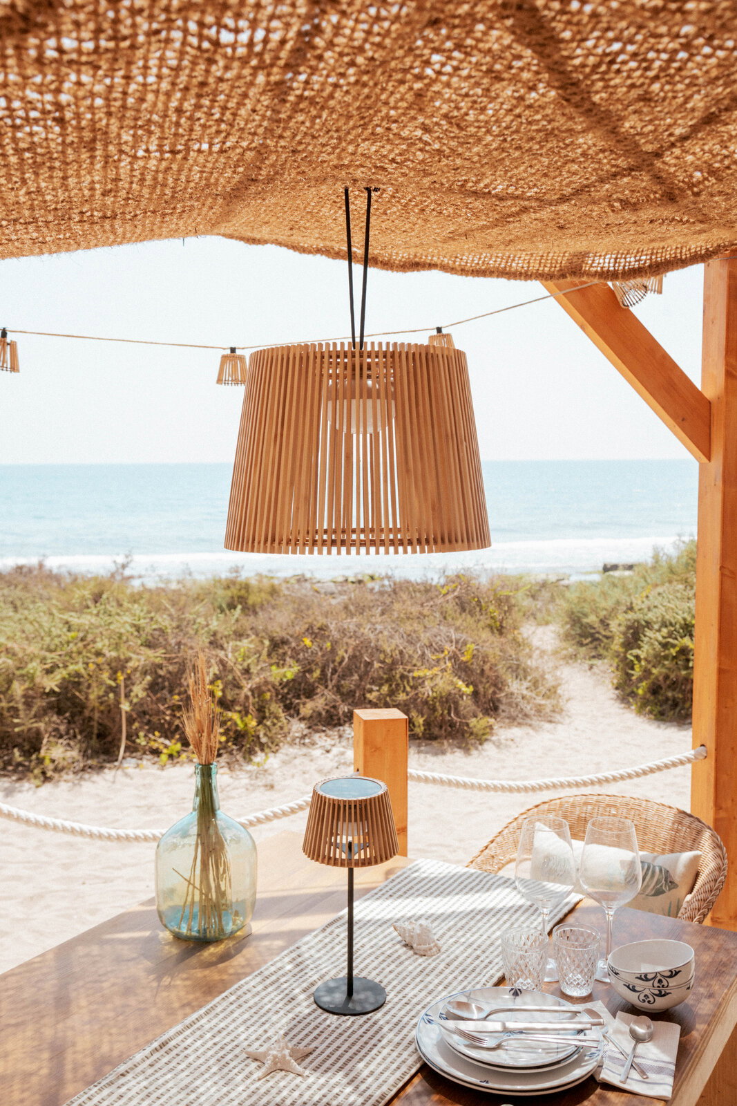 Hängelampe mit Holzlamellen unter einem geflochtenen Sonnendach über einem gedeckten Tisch am Strand, Frontansicht mit leichtem Blick von unten