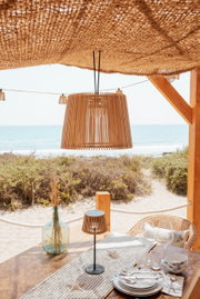 Hängelampe mit Holzlamellen unter einem geflochtenen Sonnendach über einem gedeckten Tisch am Strand, Frontansicht mit leichtem Blick von unten