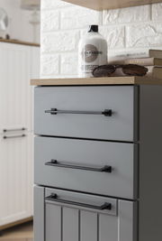 Grauer Unterschrank mit drei Schubladen, schwarzen Griffen und einer Holzplatte, aus seitlicher Perspektive fotografiert.