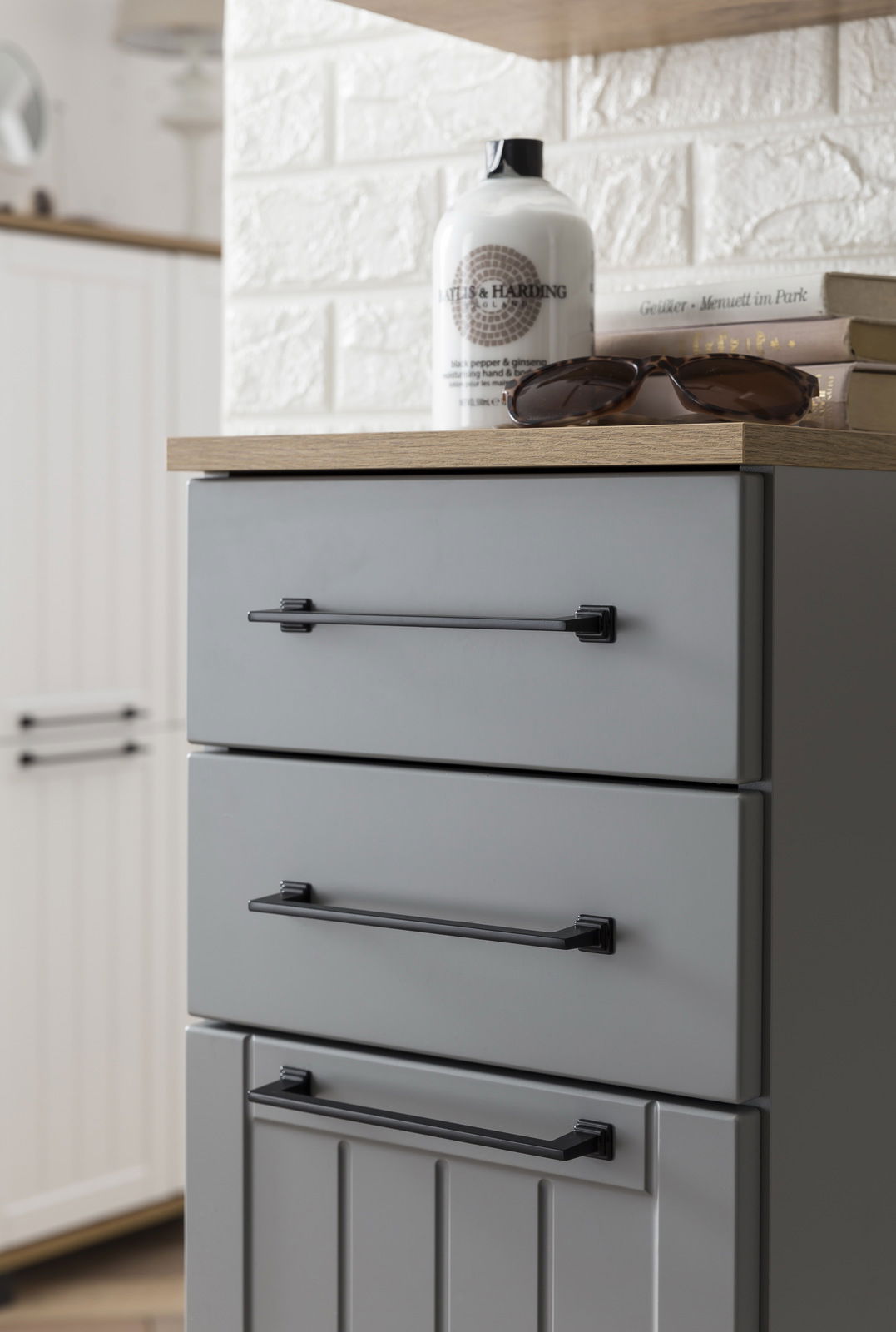 Grauer Unterschrank mit drei Schubladen, schwarzen Griffen und einer Holzplatte, aus seitlicher Perspektive fotografiert.