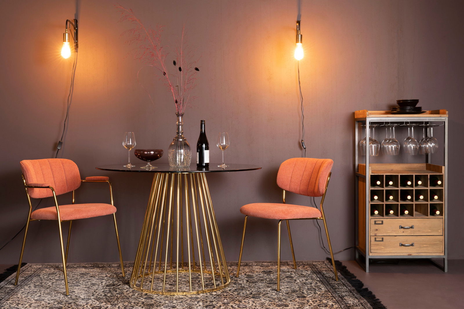 Zwei orangefarbene Stühle mit goldenen Beinen, die um einen runden Tisch mit goldener Basis in einem stilvoll beleuchteten Raum angeordnet sind, seitliche Perspektive.