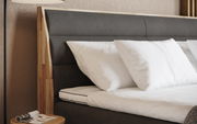 Nahaufnahme eines Boxspringbettes mit grauem Kopfteil und weißen Kissen, seitliche Perspektive