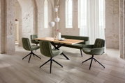 Moderner Essbereich mit rechteckigem Holzesstisch, grüner Polsterbank und vier gepolsterten Drehstühlen mit schwarzem Metallfuß in hellem Loft mit Steinwänden, Seitenansicht in leichter Schrägperspektive von vorne rechts