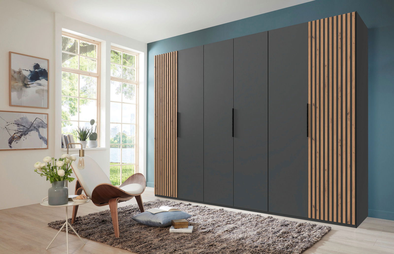 Moderner, sechstüriger Drehtürenschrank in dunklem Grau mit Holzstreifen an den Seiten, in einem hellen Raum mit großen Fenstern, von vorne betrachtet.