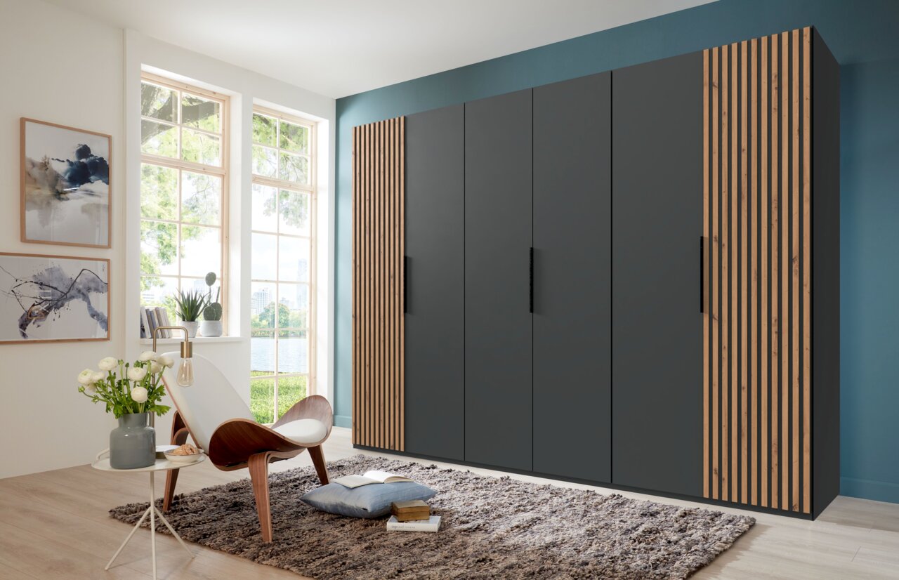 Moderner, sechstüriger Drehtürenschrank in dunklem Grau mit Holzstreifen an den Seiten, in einem hellen Raum mit großen Fenstern, von vorne betrachtet.