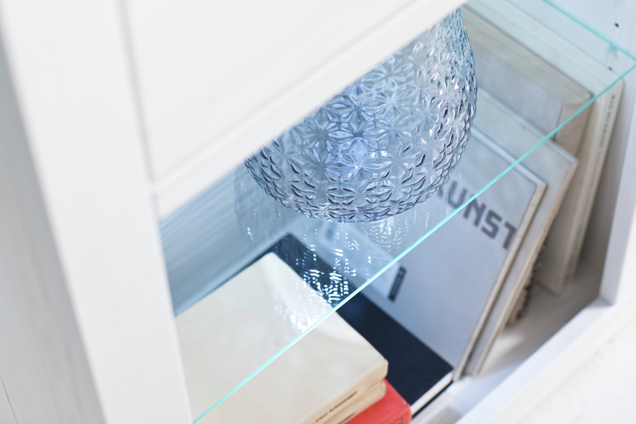 Nahaufnahme eines weißen Regals mit Glasboden, dekoriert mit einem strukturierten Glasobjekt und Büchern, von oben fotografiert.