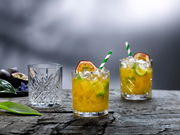 Drei Gläser auf einem Holztisch, zwei gefüllt mit einem gelben Getränk, Eis und Limettenscheiben, dekoriert mit Passionsfrucht und einem gestreiften Strohhalm, eines leer. Perspektive von vorne.