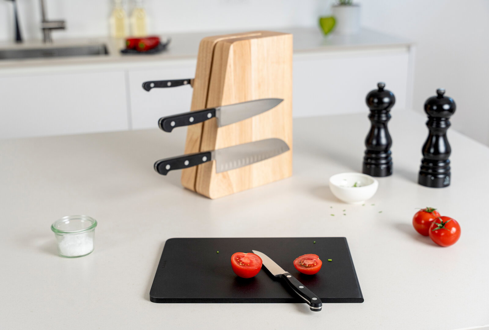 Schwarzes Schneidebrett mit halbierten Tomaten und kleinem Messer im Vordergrund, dahinter ein Holz-Messerblock mit eingesteckten Messern auf einer Küchenarbeitsplatte, Perspektive: leicht erhöhte Frontansicht.
