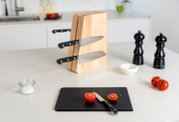 Schwarzes Schneidebrett mit halbierten Tomaten und kleinem Messer im Vordergrund, dahinter ein Holz-Messerblock mit eingesteckten Messern auf einer Küchenarbeitsplatte, Perspektive: leicht erhöhte Frontansicht.
