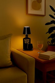 Schwarze Akku-Tischleuchte mit der Aufschrift 'Gintastic', auf einem Holztisch neben einem Sofa, mit einem Glas und Büchern, aus seitlicher Perspektive fotografiert.