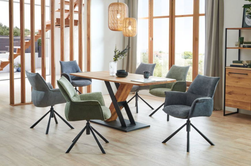 Moderner Esstisch mit Holzplatte und schwarzem Metallgestell, umgeben von gepolsterten Stühlen in verschiedenen Farben, in einem hellen Raum mit großen Fenstern, seitliche Perspektive.