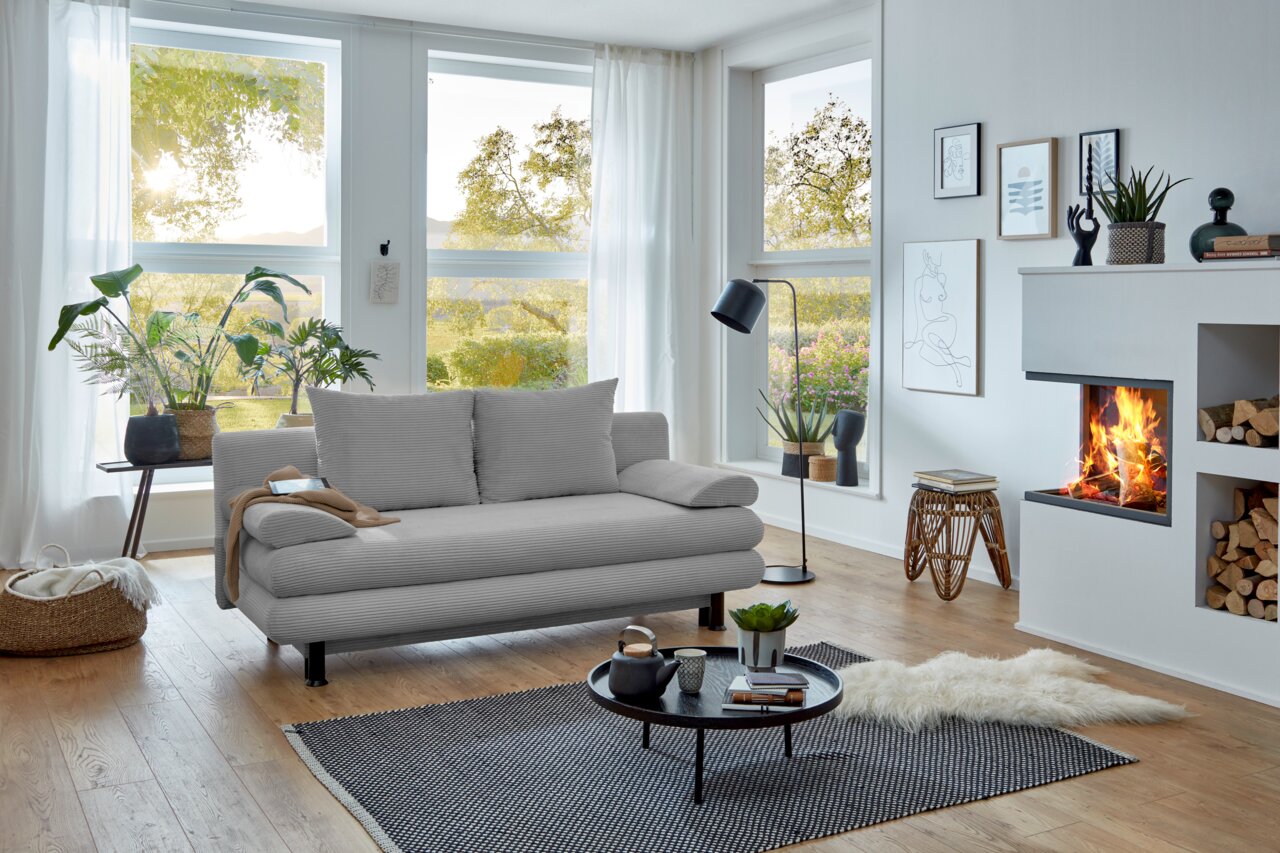 Graues Funktionssofa in modernem Wohnzimmer mit großen Fenstern, Pflanzen und Kamin, seitliche Perspektive.