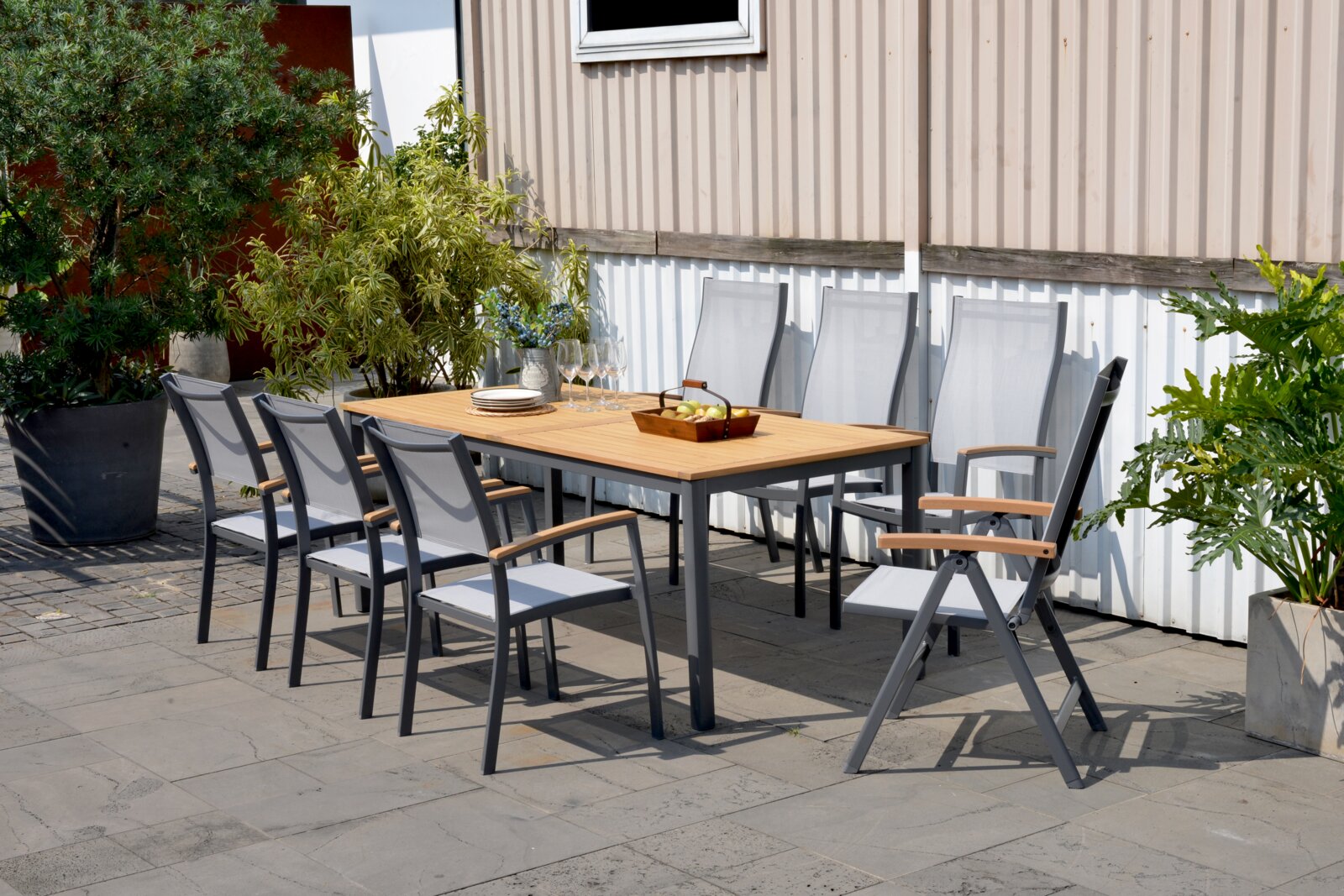 Moderner Gartentisch mit Holzplatte und Metallgestell, umgeben von acht Stühlen, auf einer Terrasse mit Pflanzen im Hintergrund, seitliche Perspektive.