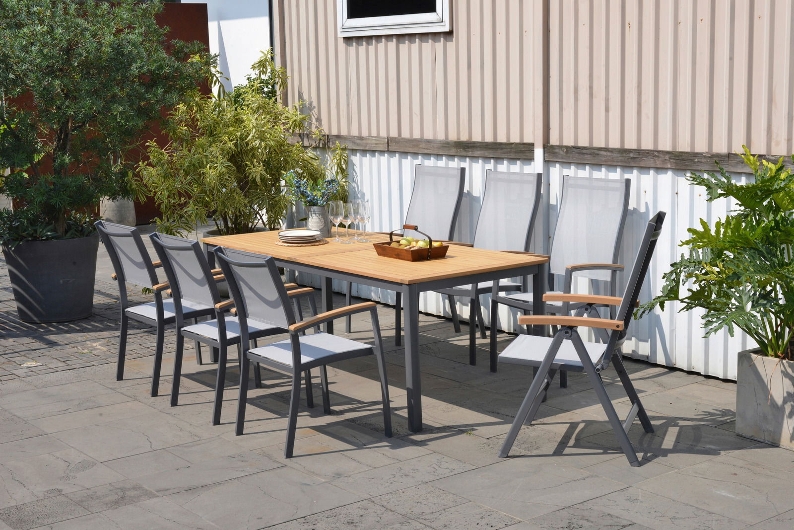 Moderner Gartentisch mit Holzplatte und Metallgestell, umgeben von acht Stühlen, auf einer Terrasse mit Pflanzen im Hintergrund, seitliche Perspektive.