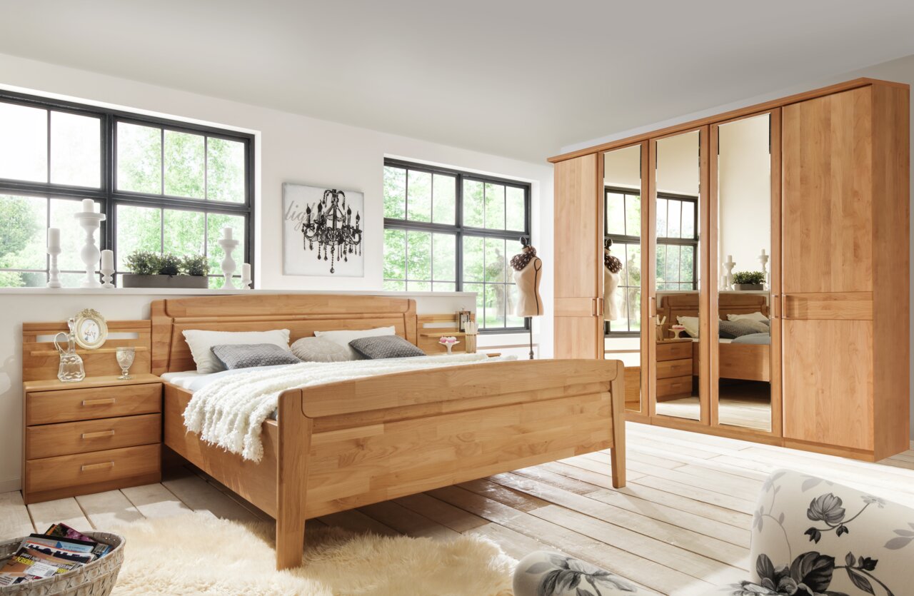 Holzbett in einem hellen Schlafzimmer mit großen Fenstern, flankiert von einem Nachttisch und einem großen Kleiderschrank mit Spiegeln, aus der Perspektive von vorne rechts.