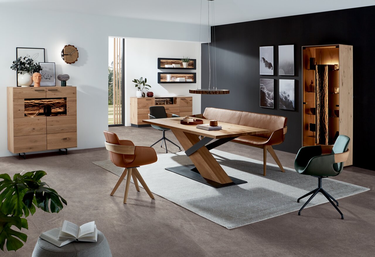 Moderner Esstisch aus Holz mit X-förmigem Gestell, umgeben von stilvollen Stühlen und einer Bank, in einem eleganten Wohnzimmer mit dunkler Akzentwand. Perspektive: seitlich.