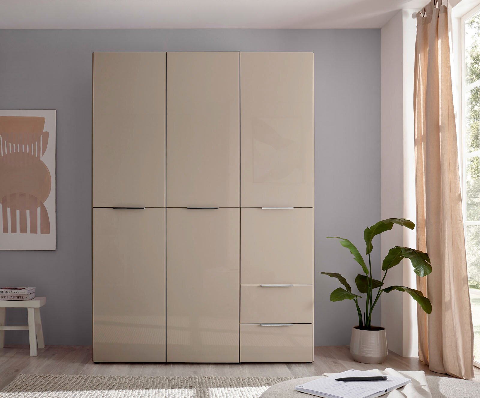 Beiger, moderner Kleiderschrank mit drei Türen und integrierten Schubladen, frontale Ansicht im hellen Schlafzimmer neben Fenster mit Vorhang und Zimmerpflanze.