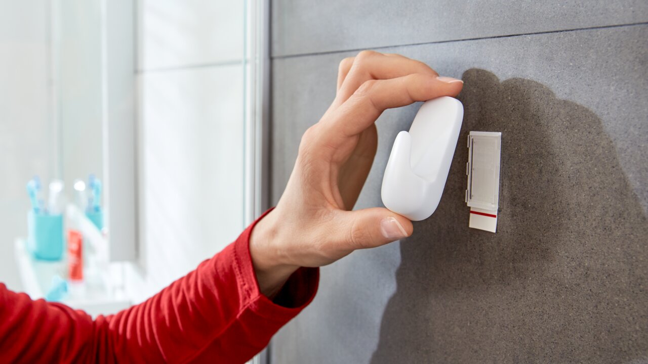 Hand befestigt einen weißen, wasserdichten tesa Powerstrip Haken an einer Badezimmerwand, seitliche Perspektive.