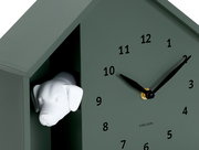 Grüne Wanduhr in Form eines Hauses mit einem weißen Hundekopf, der aus einer Öffnung herausschaut. Die Uhr hat schwarze Zeiger und Zahlen. Perspektive von schräg vorne.