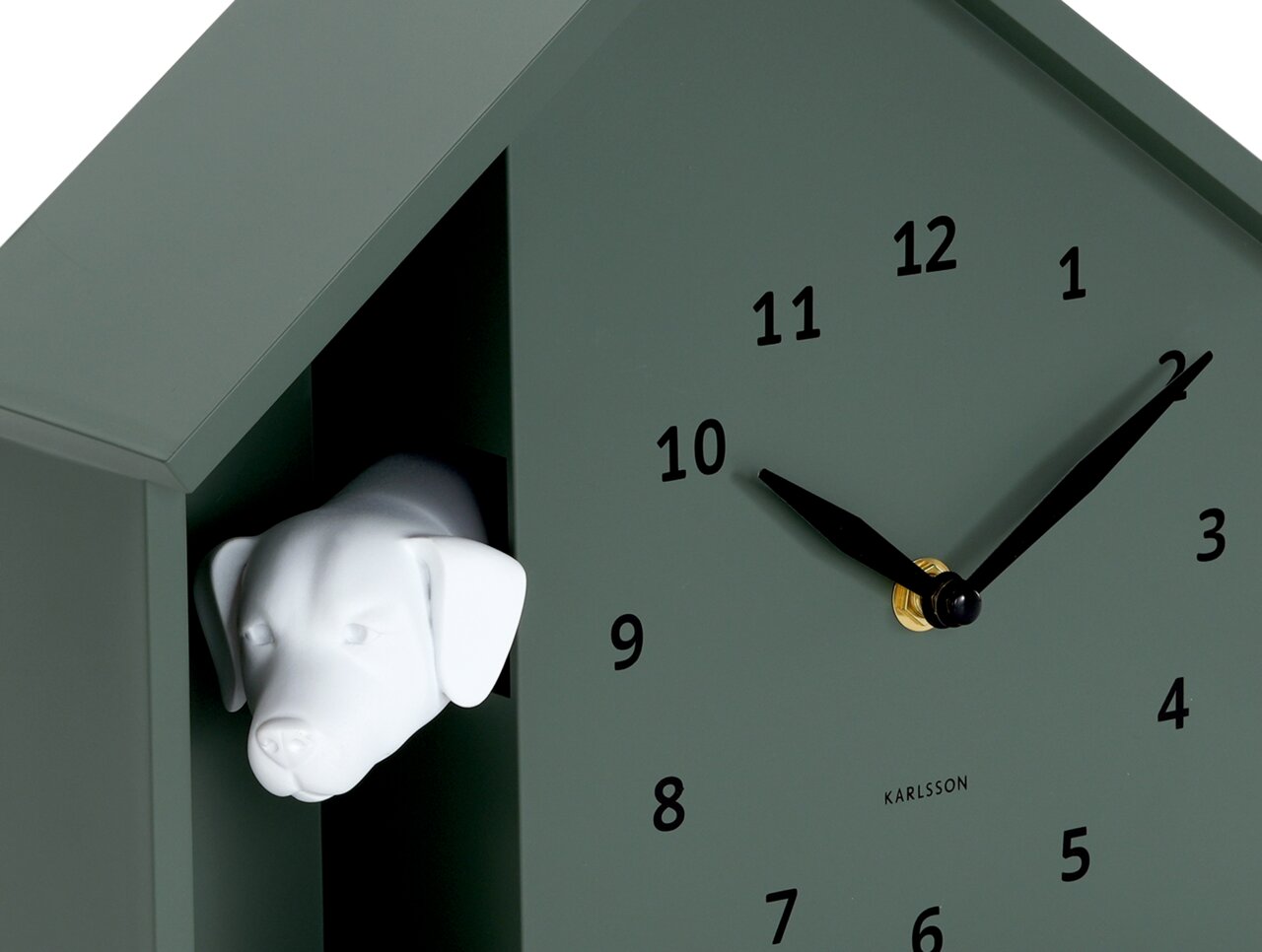 Grüne Wanduhr in Form eines Hauses mit einem weißen Hundekopf, der aus einer Öffnung herausschaut. Die Uhr hat schwarze Zeiger und Zahlen. Perspektive von schräg vorne.