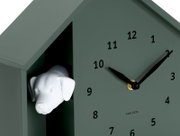 Grüne Wanduhr in Form eines Hauses mit einem weißen Hundekopf, der aus einer Öffnung herausschaut. Die Uhr hat schwarze Zeiger und Zahlen. Perspektive von schräg vorne.