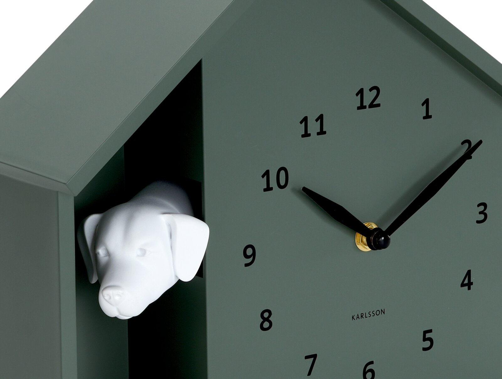 Grüne Wanduhr in Form eines Hauses mit einem weißen Hundekopf, der aus einer Öffnung herausschaut. Die Uhr hat schwarze Zeiger und Zahlen. Perspektive von schräg vorne.