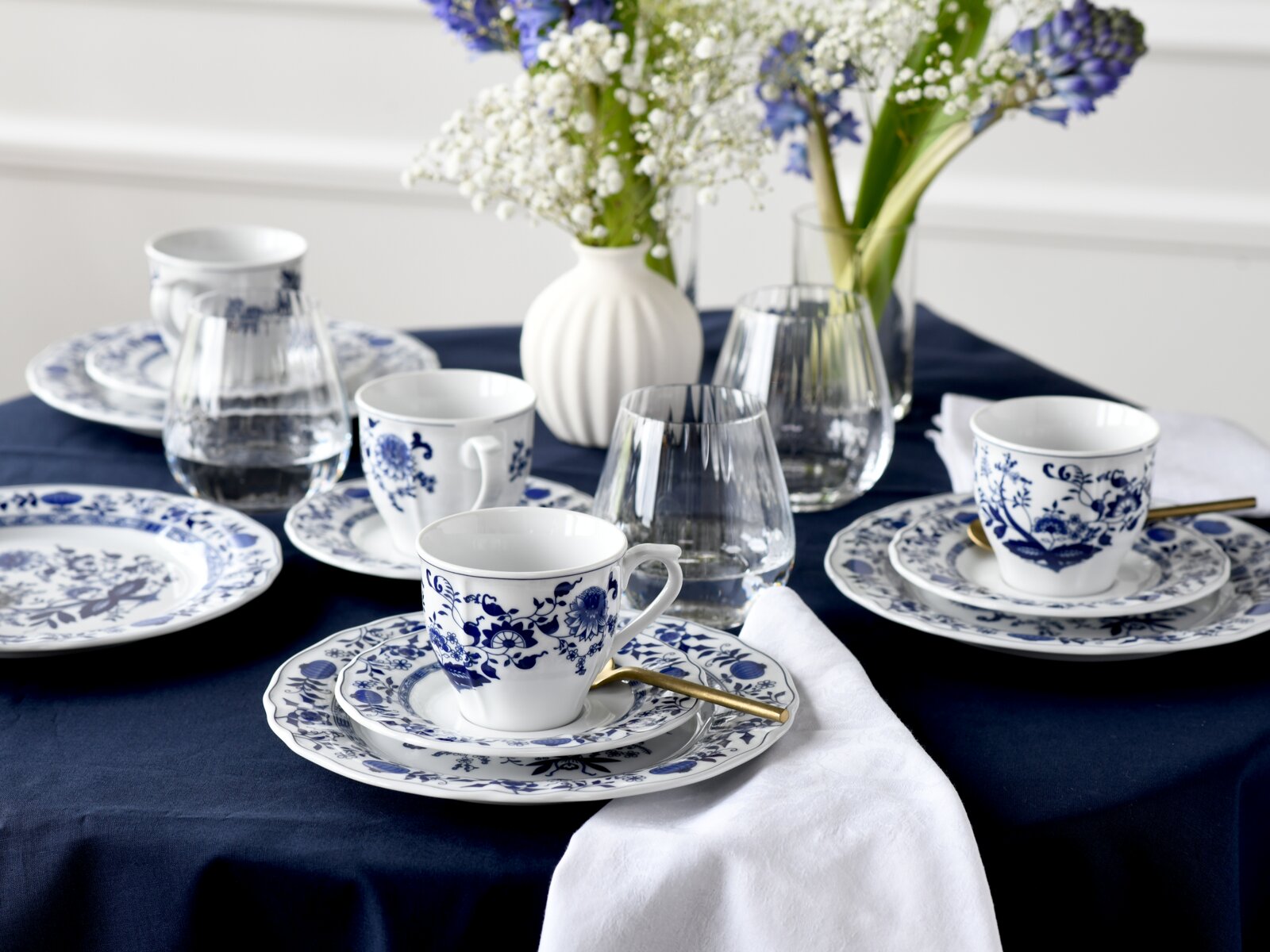 Schick gedeckter Tisch mit blau-weißem Porzellanservice (Tassen und Teller), Wassergläsern, goldfarbenem Löffel und Blumenvasen auf dunkelblauer Tischdecke, perspektivisch aus leicht erhöhter, schräger Vorderansicht aufgenommen