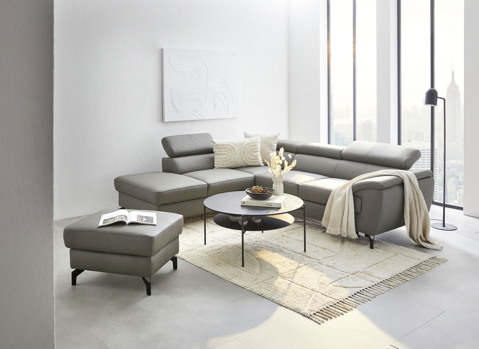 Celina Home Ecksofa FILO Moderne Eckgarnitur in einem hellen Wohnzimmer mit großen Fenstern, grauem Lederbezug und verstellbaren Kopfstützen, aus der Perspektive von vorne links fotografiert.