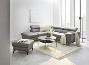 Moderne Eckgarnitur in einem hellen Wohnzimmer mit großen Fenstern, grauem Lederbezug und verstellbaren Kopfstützen, aus der Perspektive von vorne links fotografiert.