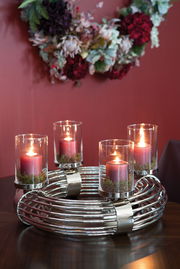 Moderner Adventskranz aus Metall mit vier brennenden roten Kerzen in Glaszylindern, auf einem Tisch vor einem roten Hintergrund, aus leicht erhöhter Perspektive.