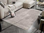 Designer Teppich in einem modernen Wohnzimmer, fotografiert aus einer leicht erhöhten Perspektive. Der Teppich hat ein dezentes Muster in Grautönen und liegt auf einem dunklen Holzboden. Im Hintergrund sind ein Sofa, ein Tisch und eine Gitarre zu sehen.