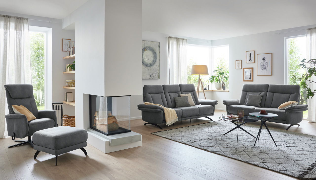 Modernes Wohnzimmer mit grauem 2,5-Sitzer Sofa, Sessel und Hocker in einer hellen, gemütlichen Umgebung. Perspektive von der Seite.