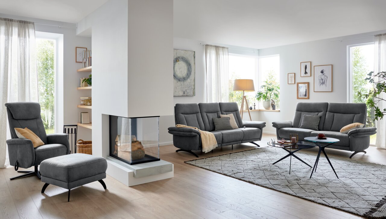 Modernes Wohnzimmer mit grauem 2,5-Sitzer Sofa, Sessel und Hocker in einer hellen, gemütlichen Umgebung. Perspektive von der Seite.
