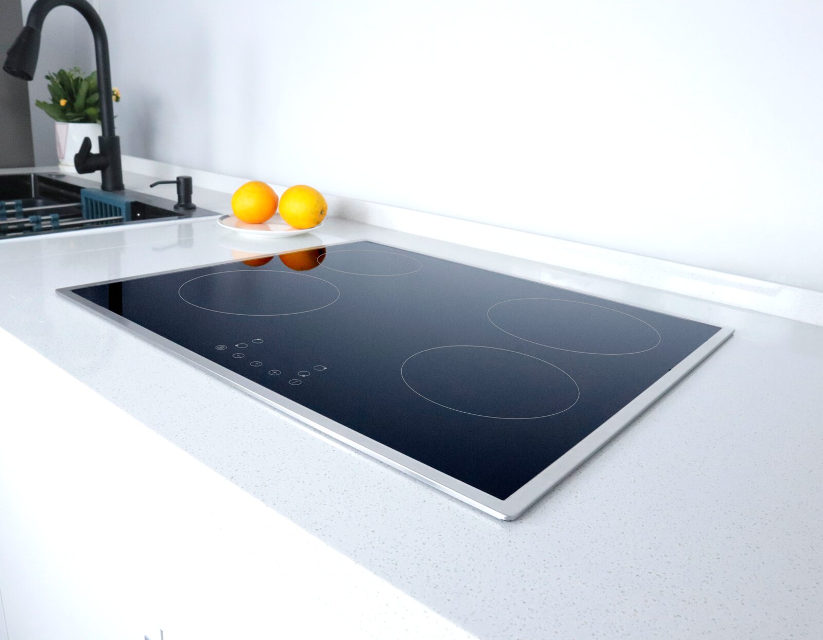 Schwarzes, flächenbündig eingebautes Glaskeramik-Kochfeld mit vier Kochzonen und Touch-Bedienfeld auf weißer Arbeitsplatte, schräg von vorne links fotografiert; im Hintergrund Spüle mit schwarzer Armatur und zwei Orangen.
