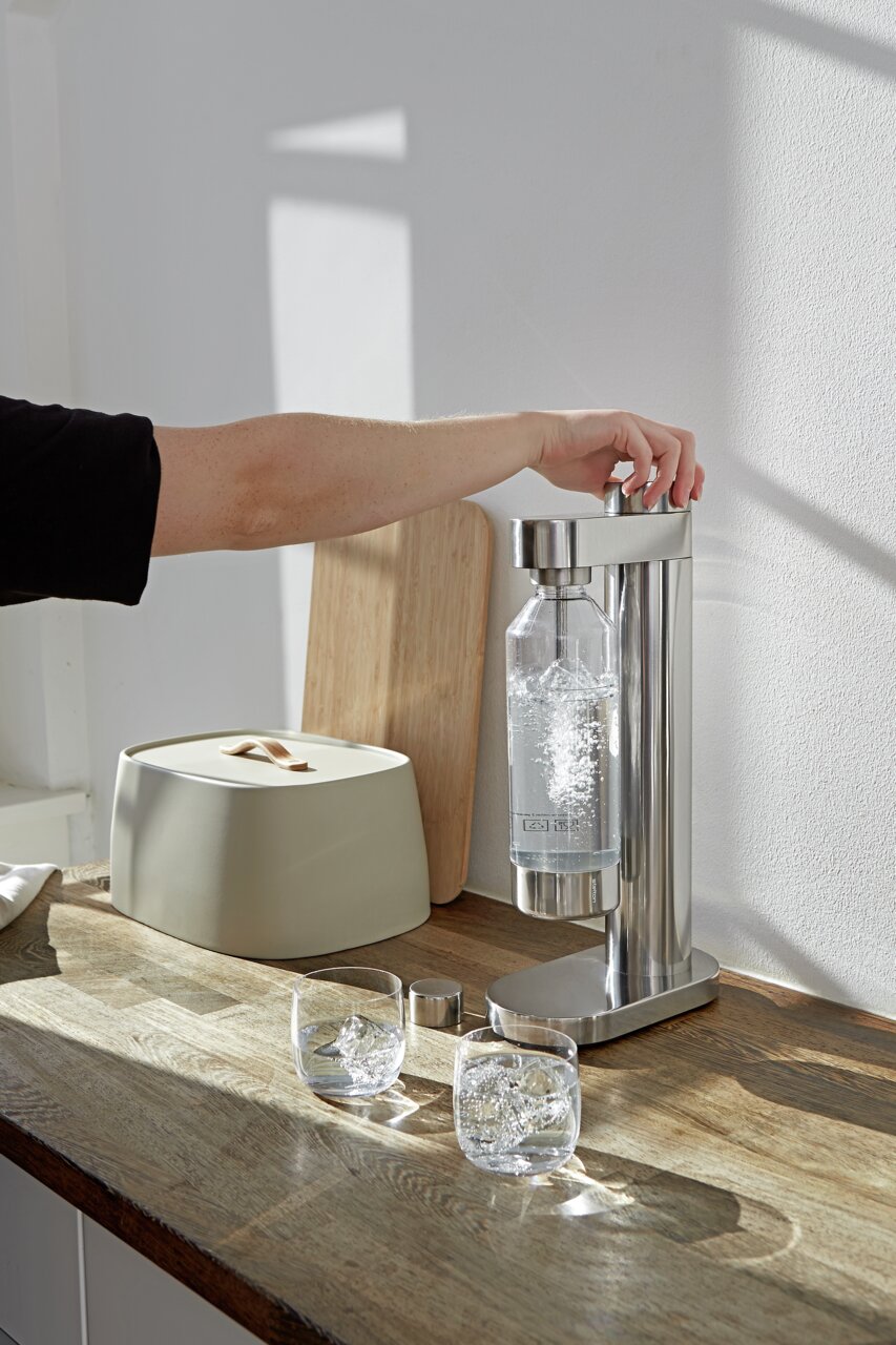 Een roestvrijstalen Brus-soda-maker op een keukenblad, gezien vanaf de zijkant, terwijl een hand het bruisproces in gang zet.